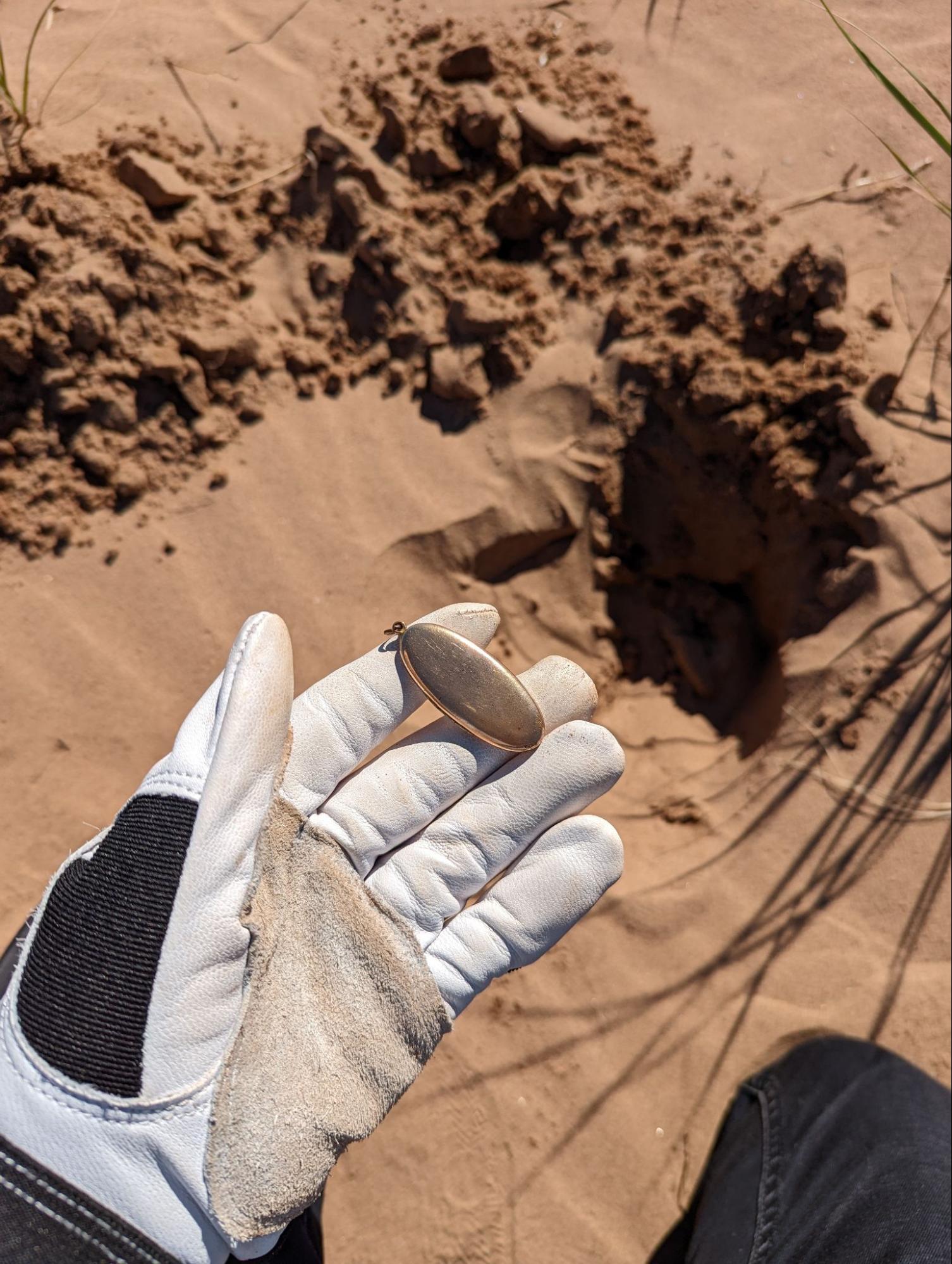 The closed heirloom locket in a gloved hand beside the freshly dug sand